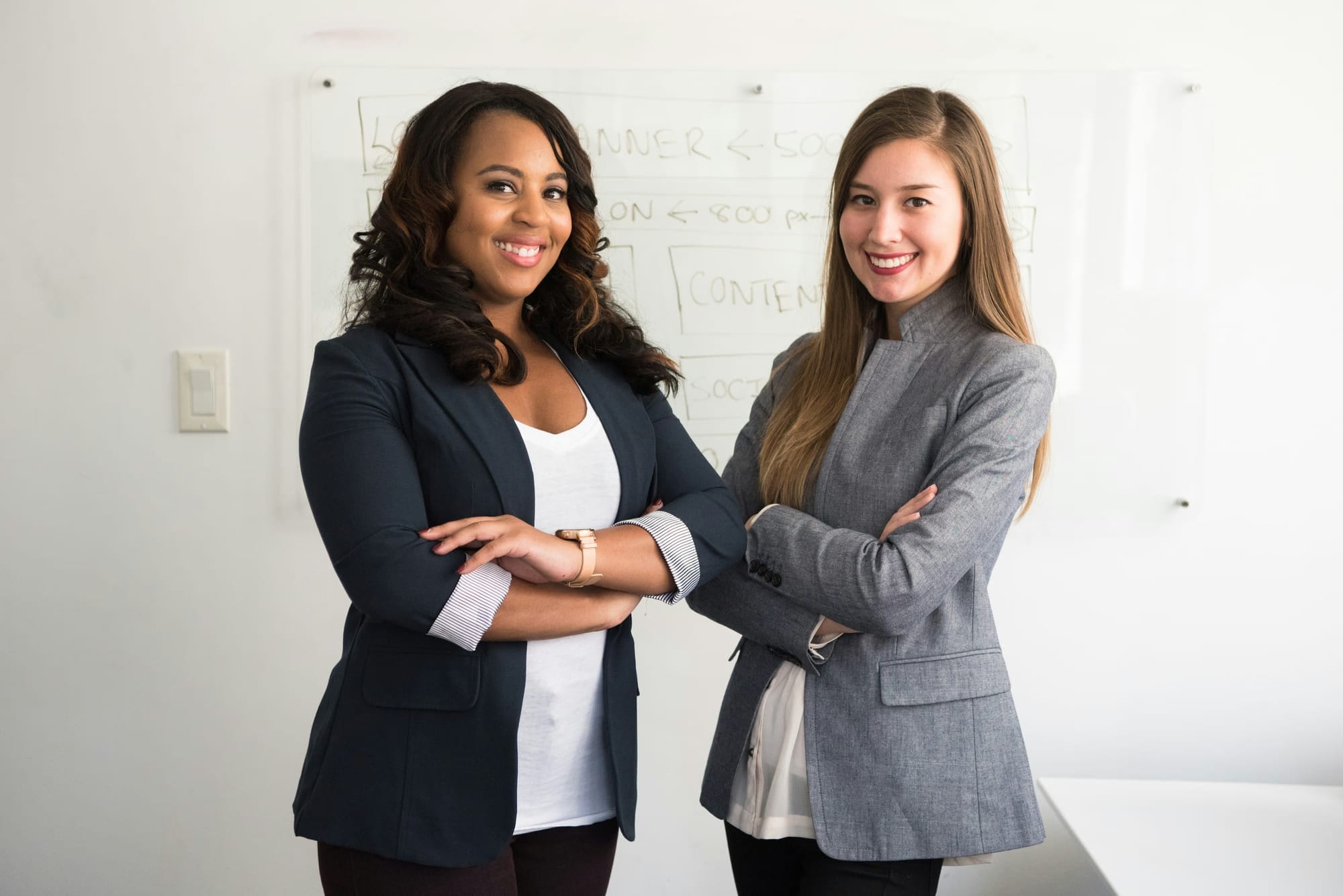 Two professional women standing side by side, confidently posing for a headshot photoshoot with a neutral background